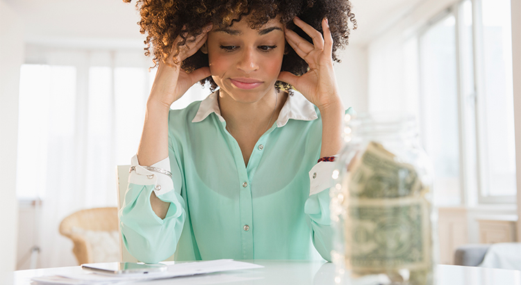 woman sits at desk head in hands looking at a paper on desk, there is a jar with dollar bills in it on desk