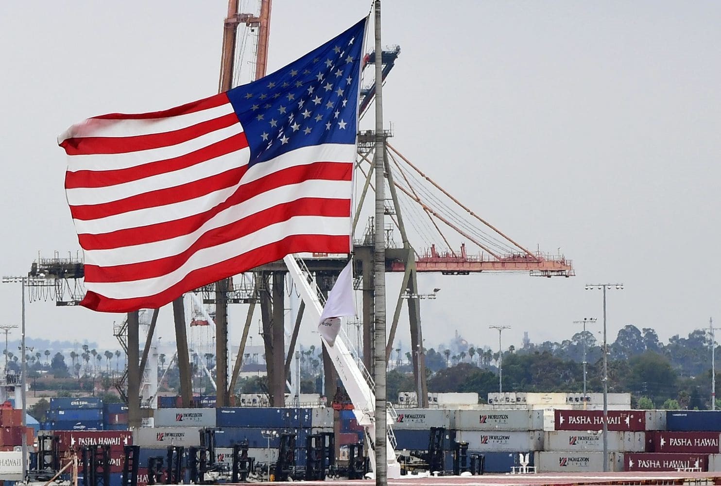 American flag flying over a shipping port with crane and shipping containers in background