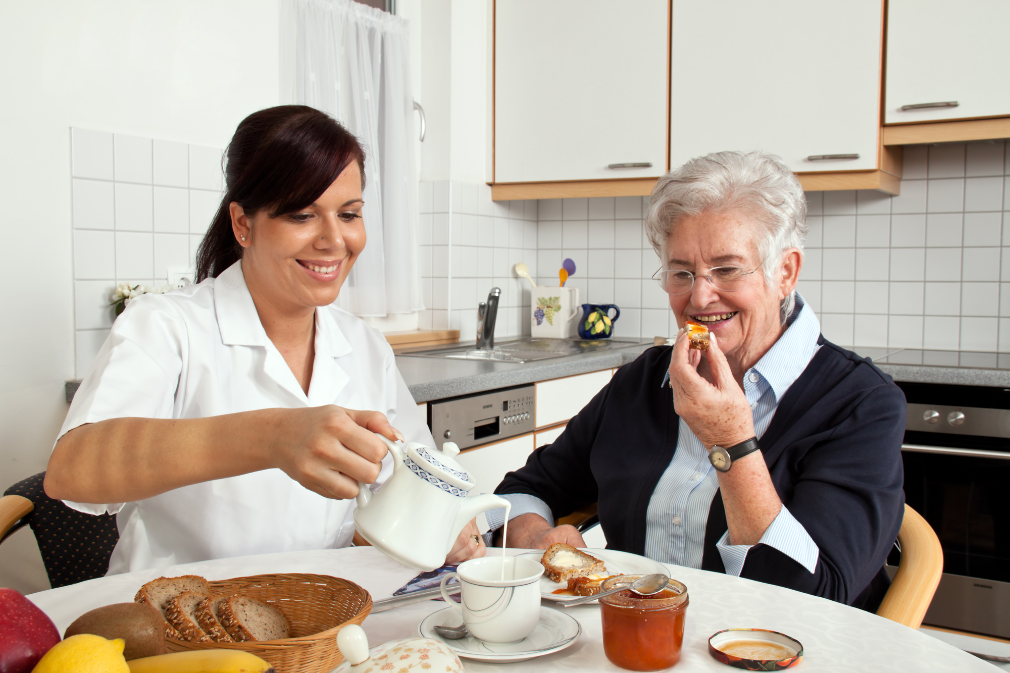 Nurse Helps Elderly Woman Two individuals sitting at a table in a home kitchen sharing breakfast; the younger person on the left pours cream into a teacup for a senior individual on the right, both smiling.