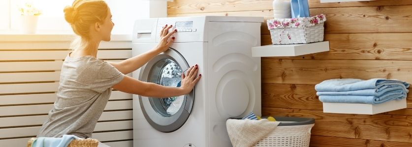 A woman kneeing on the ground starting the washing machine 