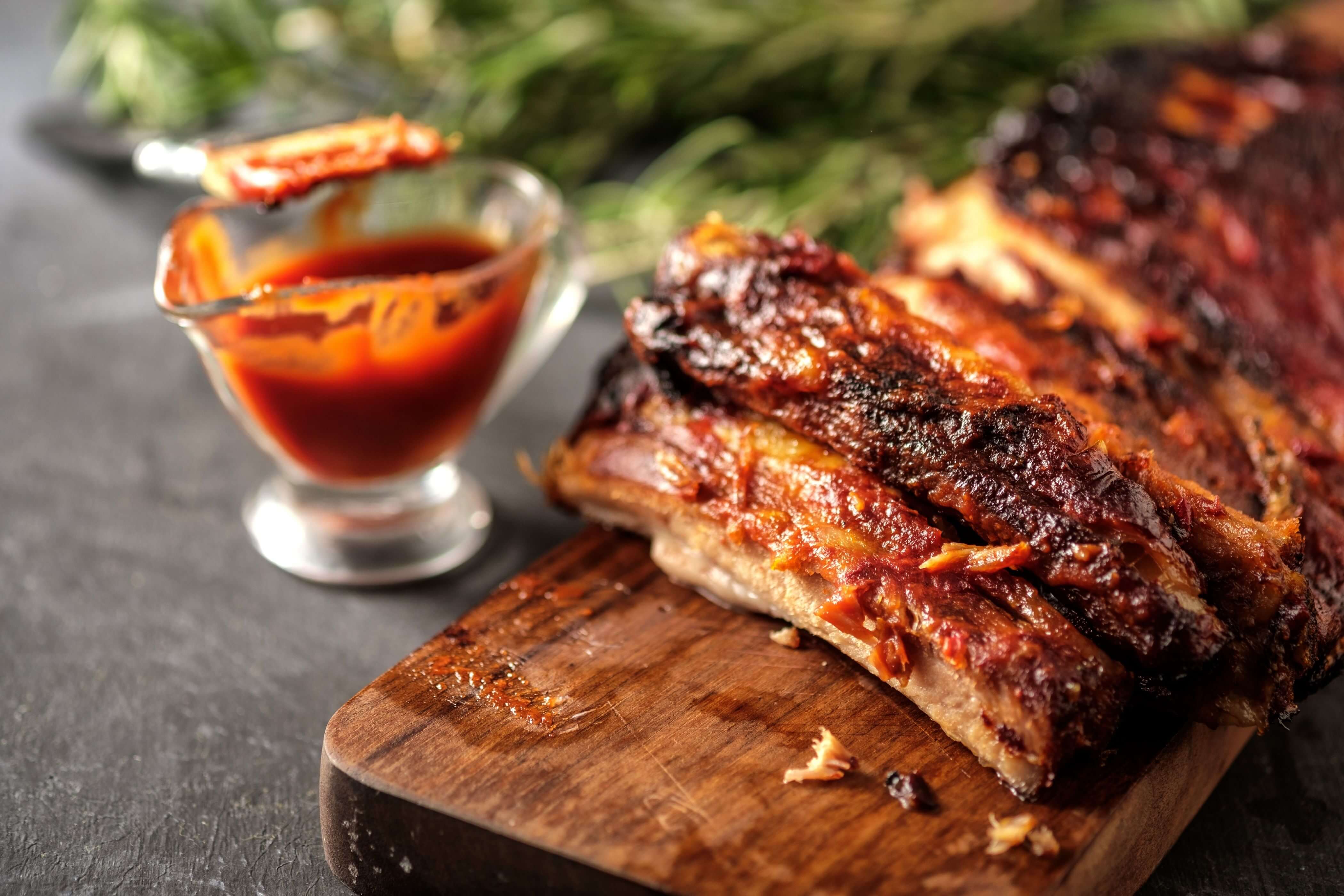 Barbecue ribs on a wooden cutting board. Glass with barbecue sauce to the left.