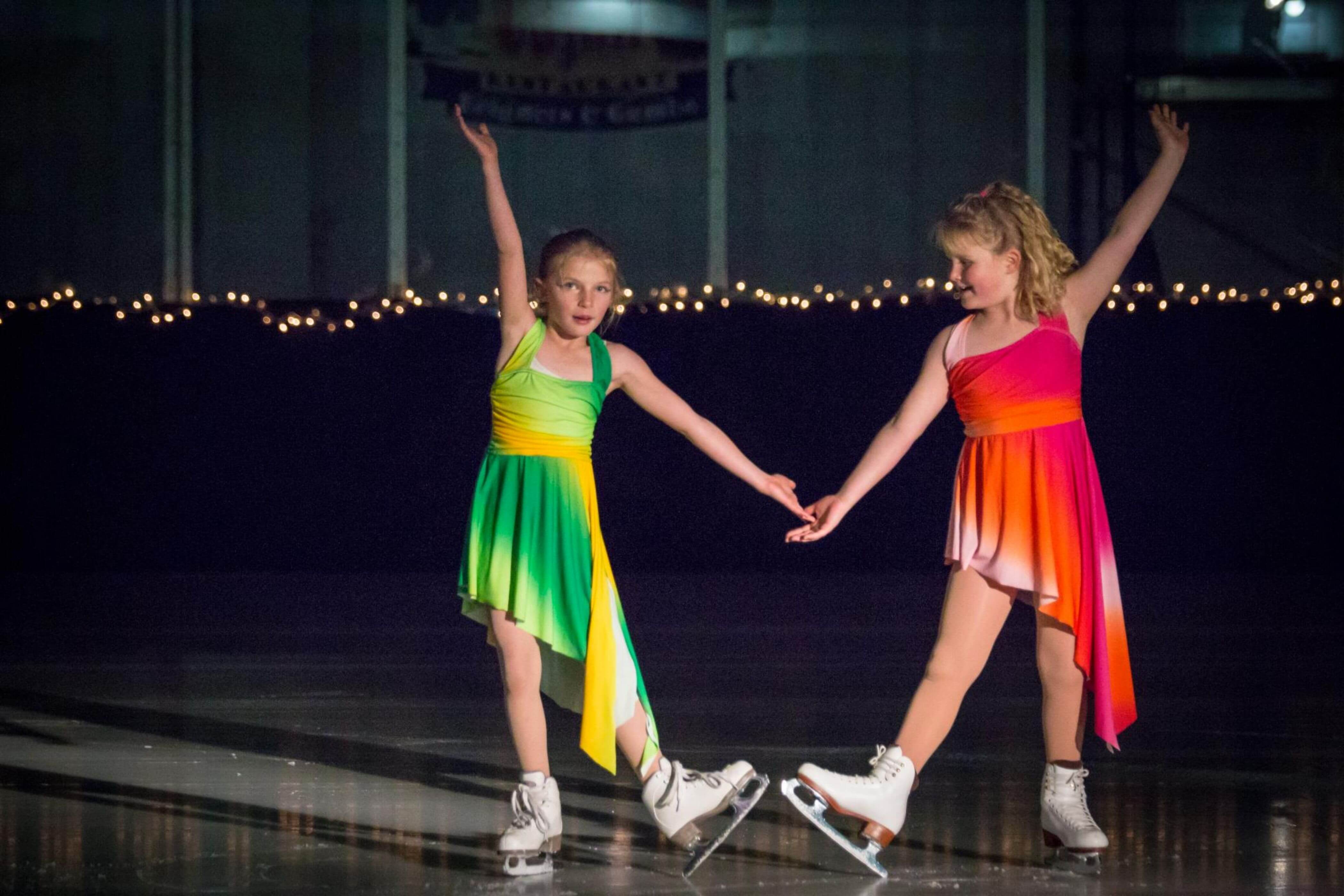 Two young female ice sakters. The left wears a yellow and green leotard. The right wears a red and yellow leotard