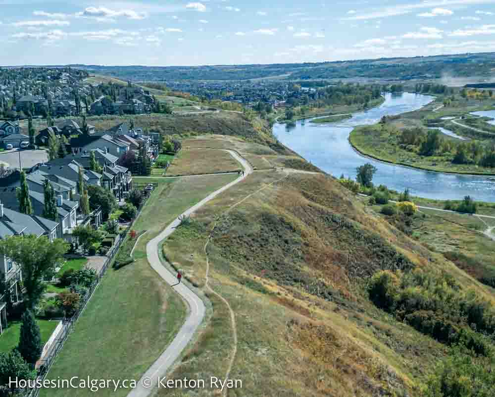 Cranston Calgary with view of the bow river valley and fish creek provincial park. There is a pathway at the top of the ridge with homes on the left and the valley on the right. The sky is blue and lots of green grass and trees in the forefront.