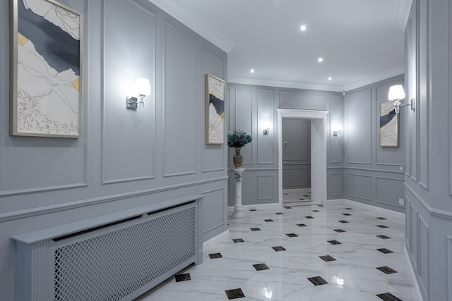 A wide and lit hallway with grey walls and white marbled tiles with black squares.