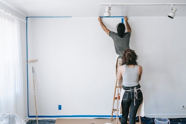 A man on a ladder putting painters tape on the edges of the ceiling, his female partner behind him holding the ladder.