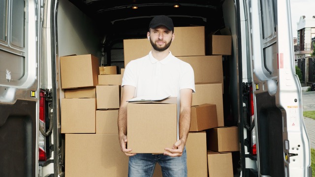 A man holding a moving box in front of a van full of moving boxes.