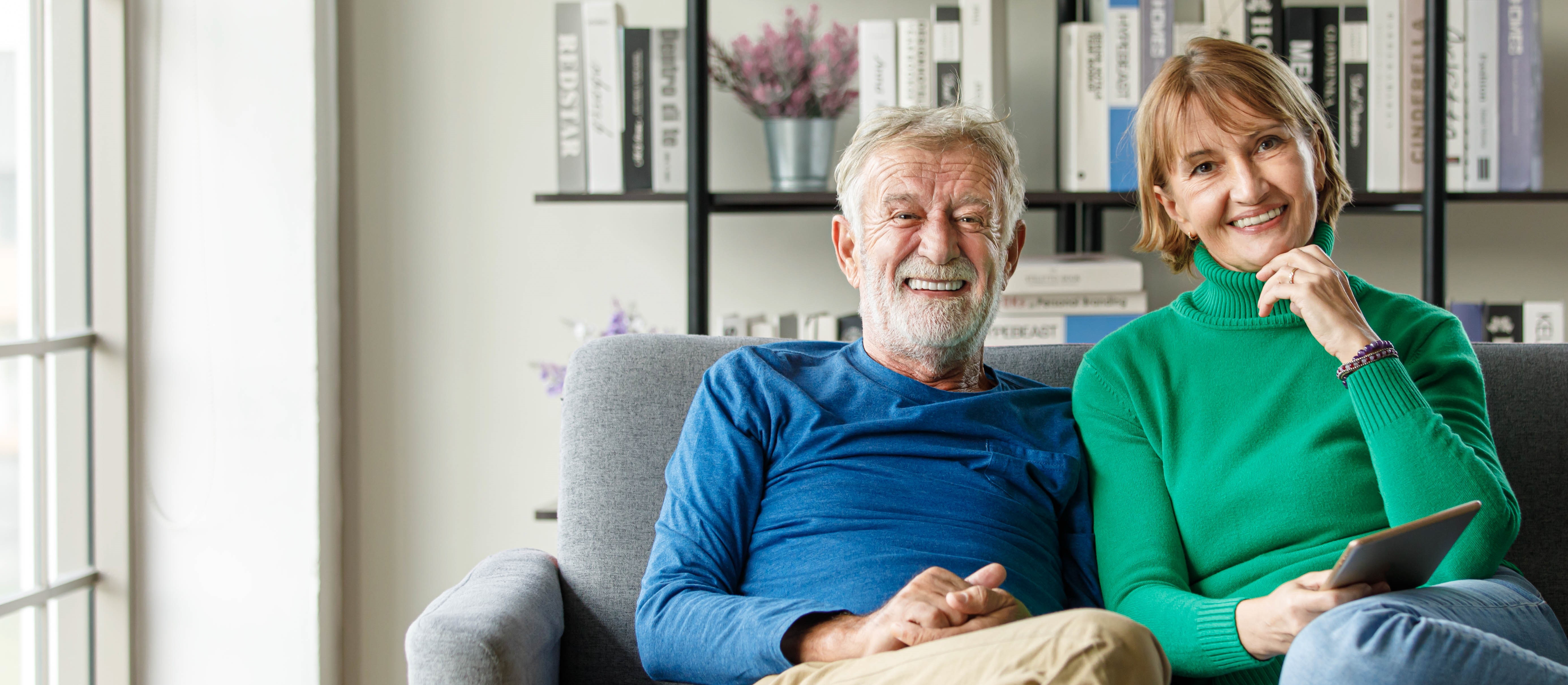 positive senior man and mature woman with tablet smiling and looking at camera while sitting on sofa in living
