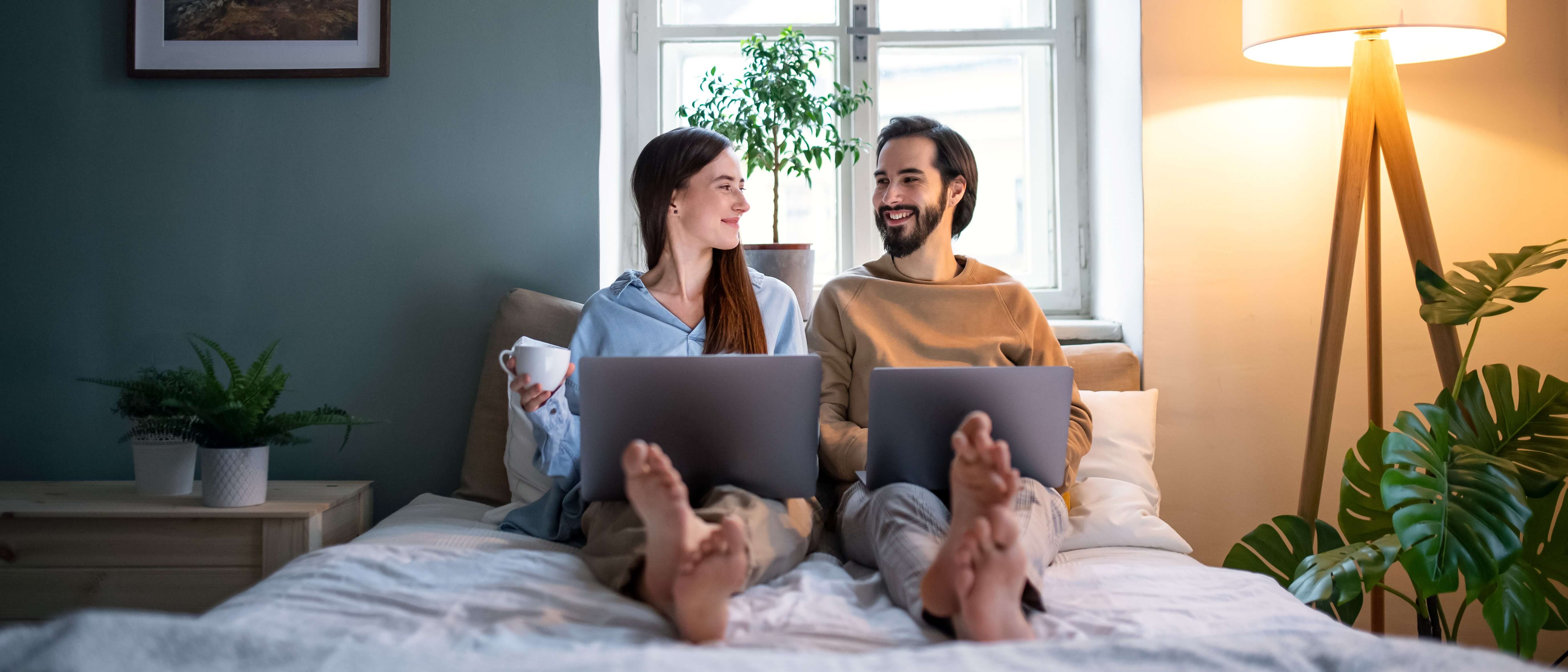 Young couple using laptops in bed to view virtual home tours